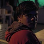 Homem com capuz vermelho em ambiente noturno, possivelmente em um evento cultural.