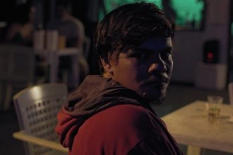 Homem com capuz vermelho em ambiente noturno, possivelmente em um evento cultural.