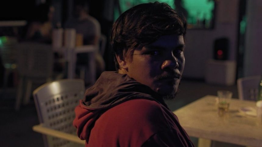 Homem com capuz vermelho em ambiente noturno, possivelmente em um evento cultural.