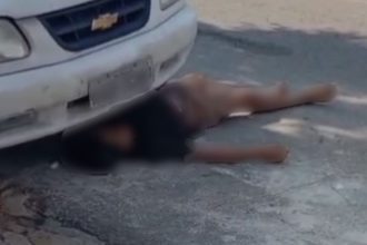 Homem deitado no chão perto de carro, fingindo estar morto para escapar após roubo.