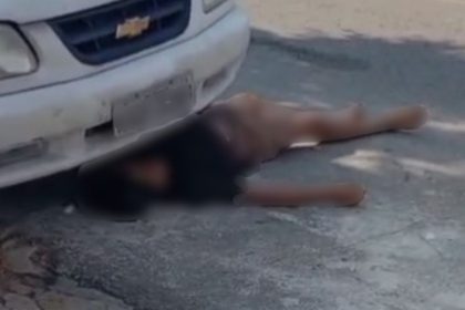 Homem deitado no chão perto de carro, fingindo estar morto para escapar após roubo.