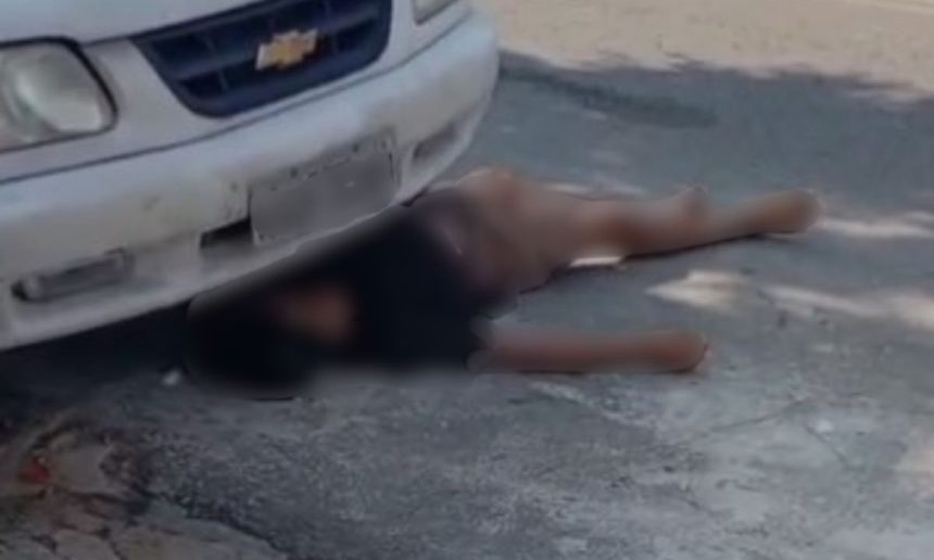 Homem deitado no chão perto de carro, fingindo estar morto para escapar após roubo.