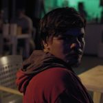 Homem em bar com iluminação noturna, olhar direto para a câmera.