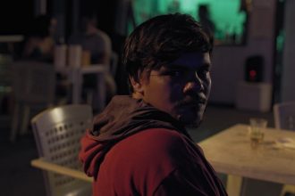 Homem em bar com iluminação noturna, olhar direto para a câmera.