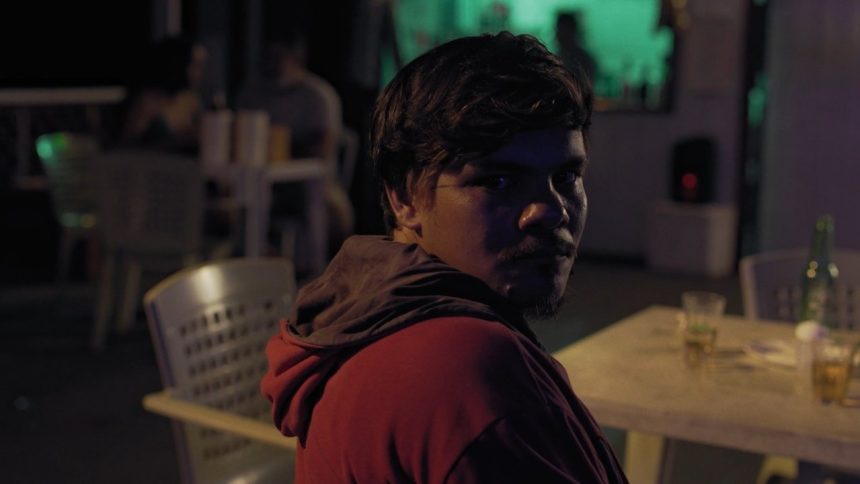 Homem em bar com iluminação noturna, olhar direto para a câmera.