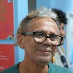Homem sorrindo com óculos em evento de arte na Casa das Artes.