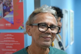 Homem sorrindo com óculos em evento de arte na Casa das Artes.