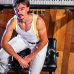 Homem vestido como Freddie Mercury em frente a um piano.
