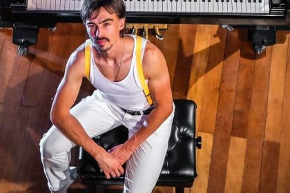 Homem vestido como Freddie Mercury em frente a um piano.
