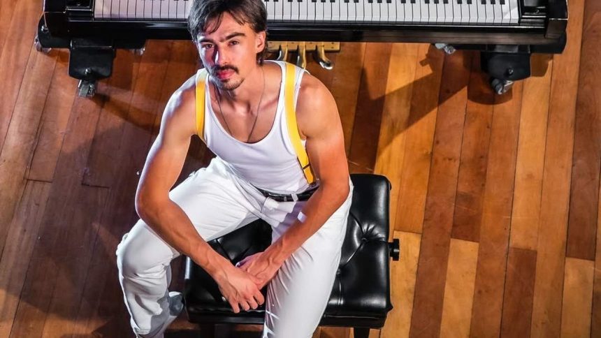 Homem vestido como Freddie Mercury em frente a um piano.
