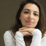 Mulher de meia-idade sorrindo, com as mãos sob o queixo, vestindo camisa branca.