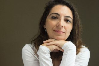 Mulher de meia-idade sorrindo, com as mãos sob o queixo, vestindo camisa branca.