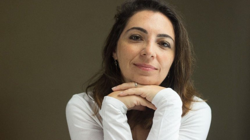 Mulher de meia-idade sorrindo, com as mãos sob o queixo, vestindo camisa branca.