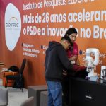 Pessoas observando robôs em um estande do Instituto Eldorado, com logotipo em destaque. Foco em inovação e tecnologia.