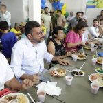 Pessoas reunidas em um evento social comendo em mesas longas, com alimentos servidos em pratos.