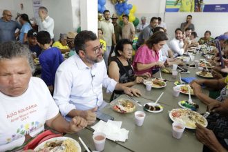 Pessoas reunidas em um evento social comendo em mesas longas, com alimentos servidos em pratos.