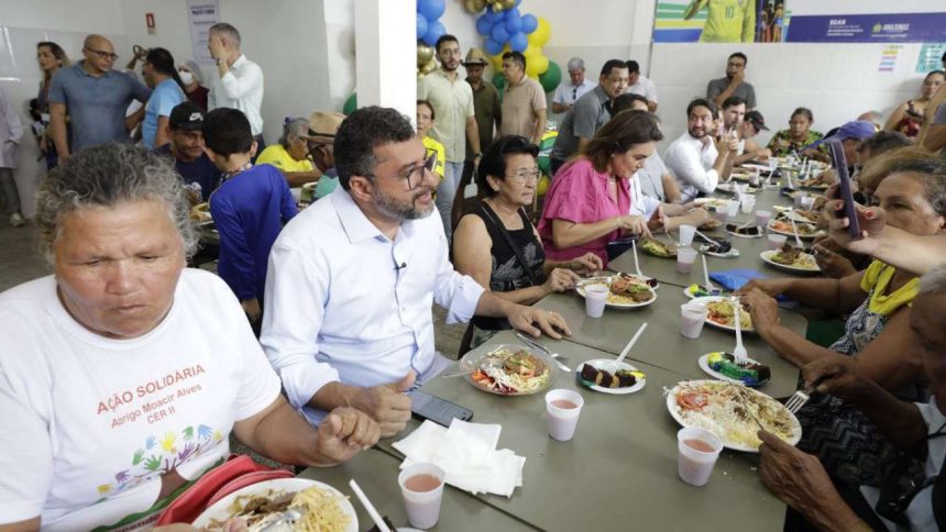 Pessoas reunidas em um evento social comendo em mesas longas, com alimentos servidos em pratos.
