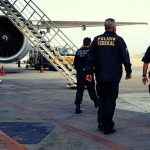Policiais federais em ação em um aeroporto, com avião ao fundo.