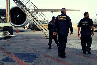 Policiais federais em ação em um aeroporto, com avião ao fundo.