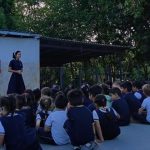 Professora fala para alunos sentados no chão em evento escolar.