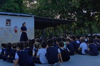Professora fala para alunos sentados no chão em evento escolar.