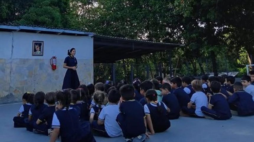 Professora fala para alunos sentados no chão em evento escolar.