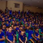 Público em evento cultural infantil, com crianças vestindo uniformes escolares em auditório.