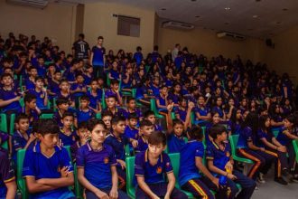 Público em evento cultural infantil, com crianças vestindo uniformes escolares em auditório.