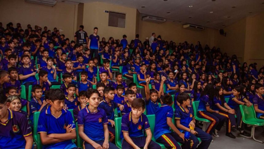 Público em evento cultural infantil, com crianças vestindo uniformes escolares em auditório.