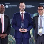 Três homens em trajes formais em um evento, um deles segura um prêmio.