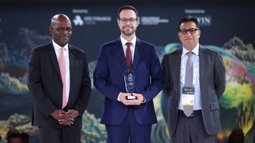 Três homens em trajes formais em um evento, um deles segura um prêmio.