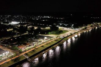 Vista aérea noturna de Maués com iluminação pública em LED, refletindo na água.
