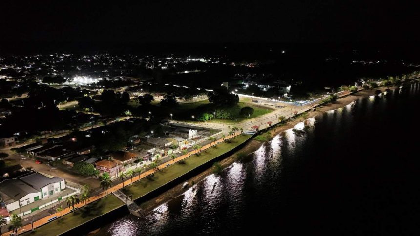 Vista aérea noturna de Maués com iluminação pública em LED, refletindo na água.