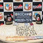 Apreensão de drogas: diversos pacotes e dinheiro sobre uma mesa, com emblemas da Rocam ao fundo.