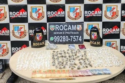 Apreensão de drogas: diversos pacotes e dinheiro sobre uma mesa, com emblemas da Rocam ao fundo.