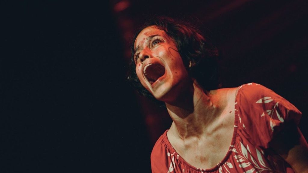 Atriz em performance teatral intensa, com expressão de sofrimento e sangue no rosto