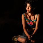 Atriz sentada com roupa colorida e colar em cena da peça 'Eu Capitu'