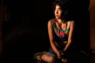 Atriz sentada com roupa colorida e colar em cena da peça 'Eu Capitu'