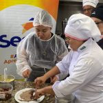 Aula de gastronomia com pessoas em uniformes de chef preparando alimentos, com ingredientes na bancada.