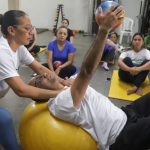Aula de pilates com instrutora auxiliando um aluno em exercício.