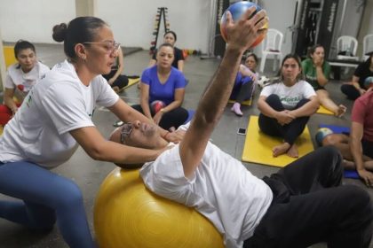 Aula de pilates com instrutora auxiliando um aluno em exercício.