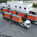 Caminhões transportando geradores laranja em um pátio industrial.