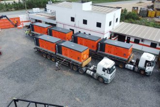 Caminhões transportando geradores laranja em um pátio industrial.