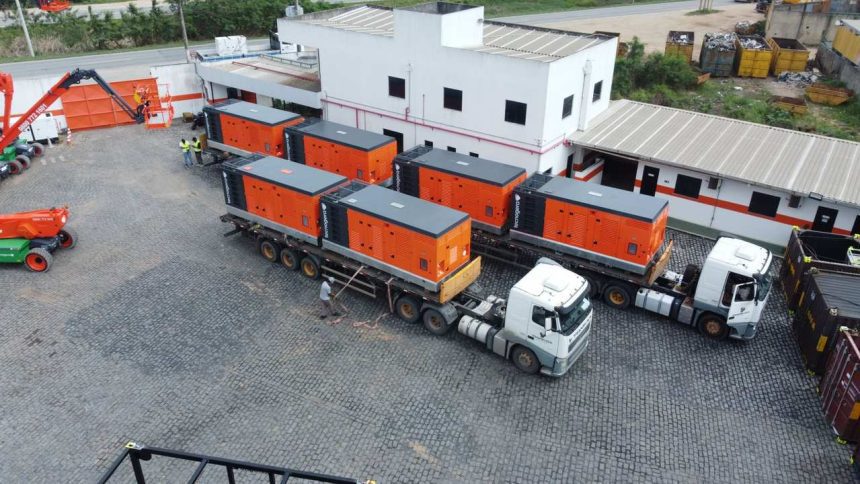 Caminhões transportando geradores laranja em um pátio industrial.