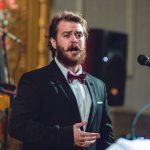 Cantor de terno e gravata borboleta se apresenta com microfone.