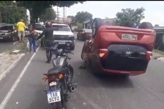 Carro capotado na rua após acidente com roda solta. Motocicleta e pessoas ao redor.