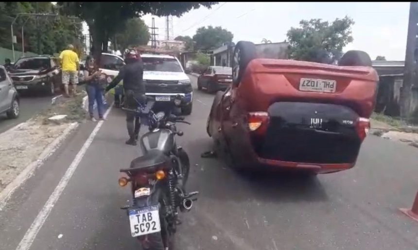 Carro capotado na rua após acidente com roda solta. Motocicleta e pessoas ao redor.