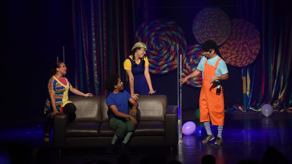 Cena de espetáculo teatral infantil com figurinos coloridos e cenografia lúdica.