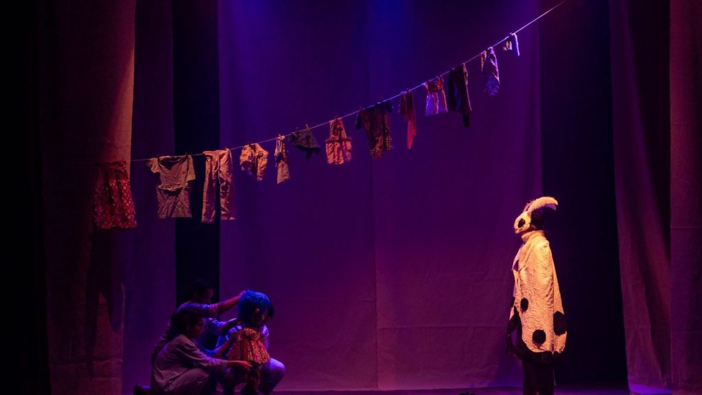 Cena de peça teatral: figurino de animal e roupas penduradas em varal.