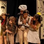 Cena teatral com atores vestidos com figurinos de época e cenários desenhados.